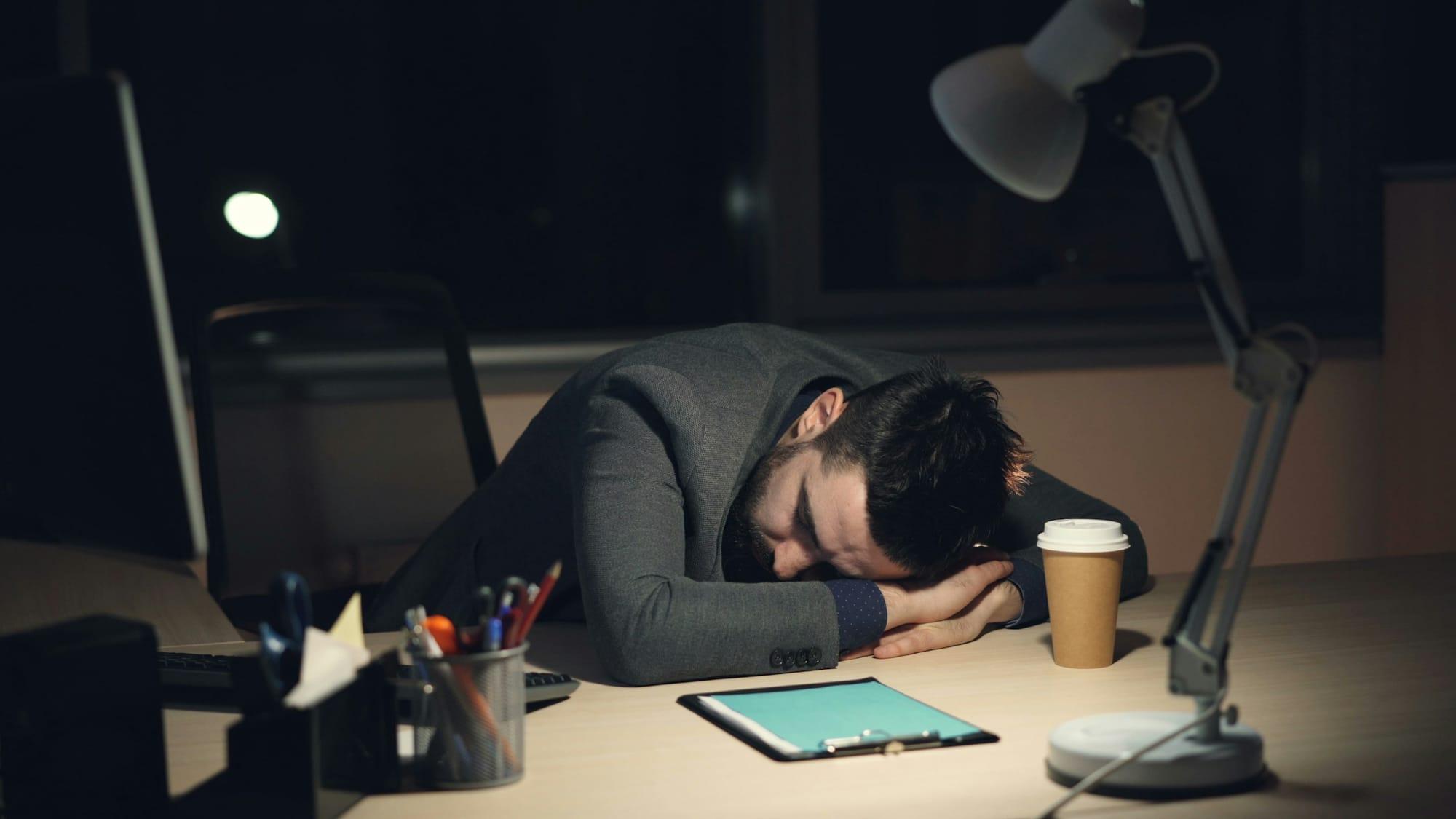 Man asleep at his desk late at night, beside a coffee cup and a tablet